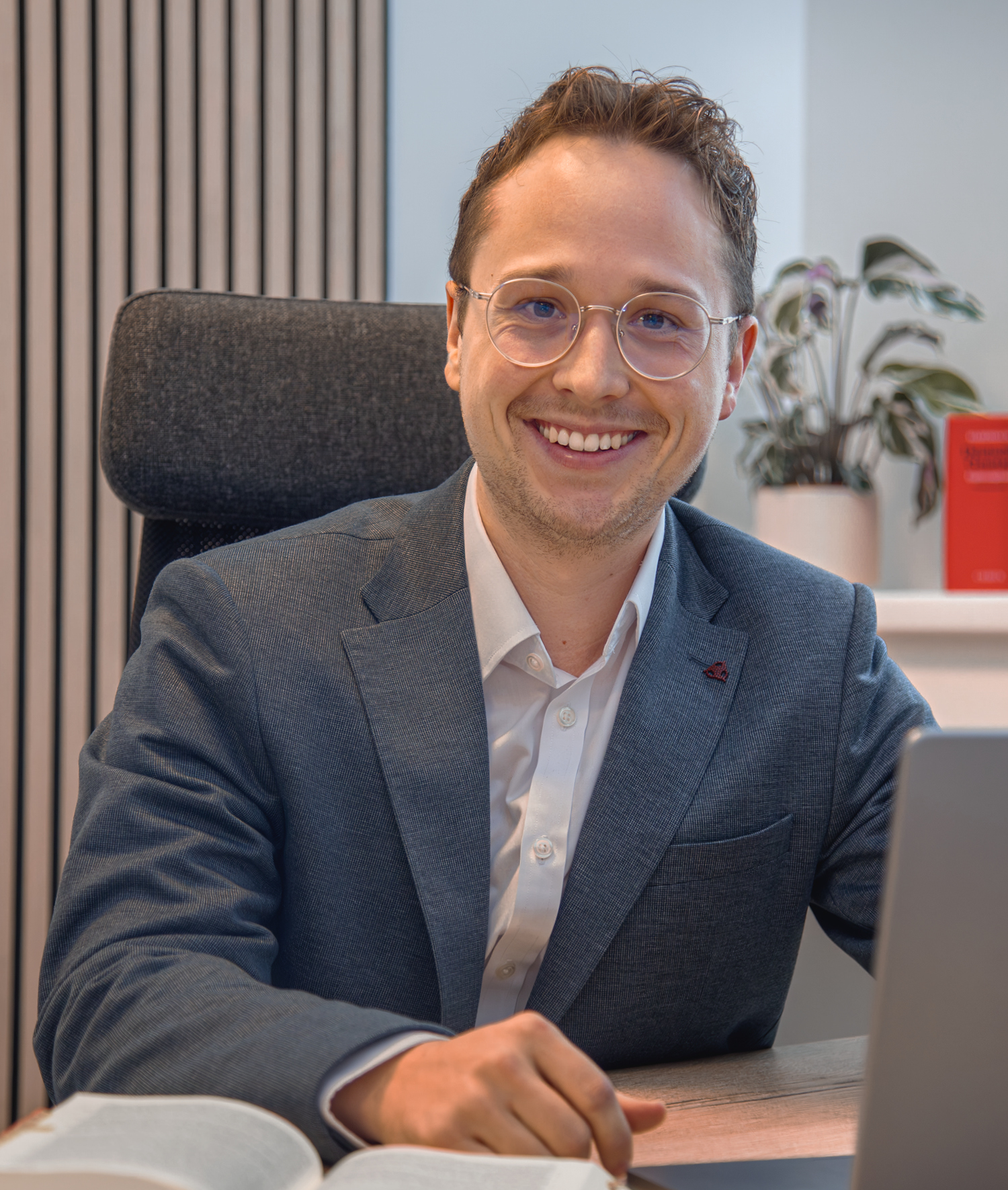 Porträt eines lächelnden Manuel Zettl mit Brille am Schreibtisch vor Laptop und aufgeschlagenem Buch.